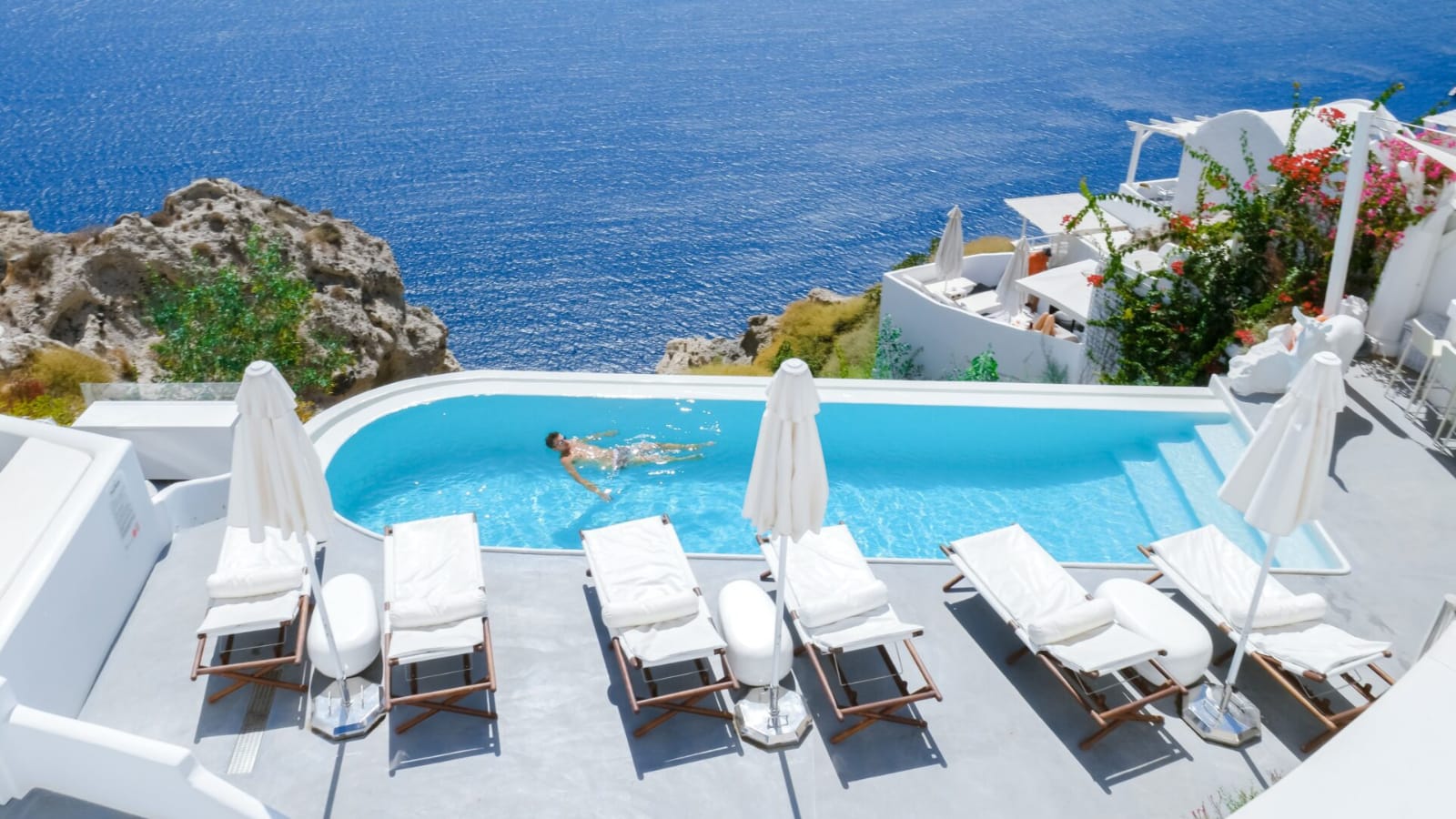 Man floating alone in an azure infinity pool at a luxury resort on a Greek island, likely Santorini, with the pool set at the edge of a dramatic coastal cliff overlooking the sea