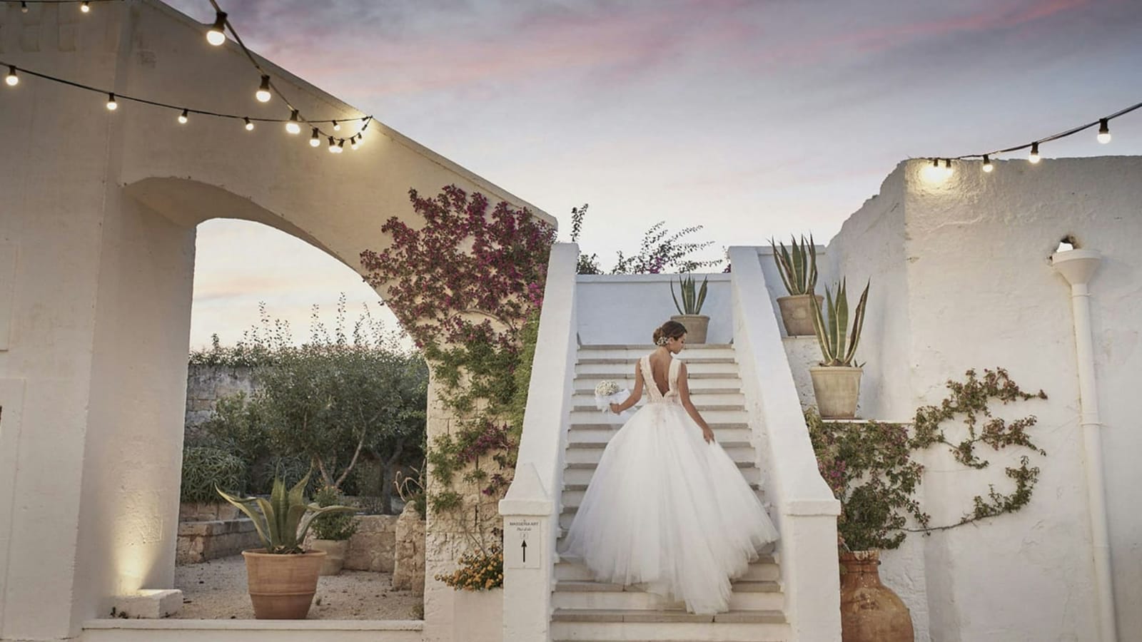 A beautiful bride walking up the stairs beneath a sunset sky, holding a bouquet.