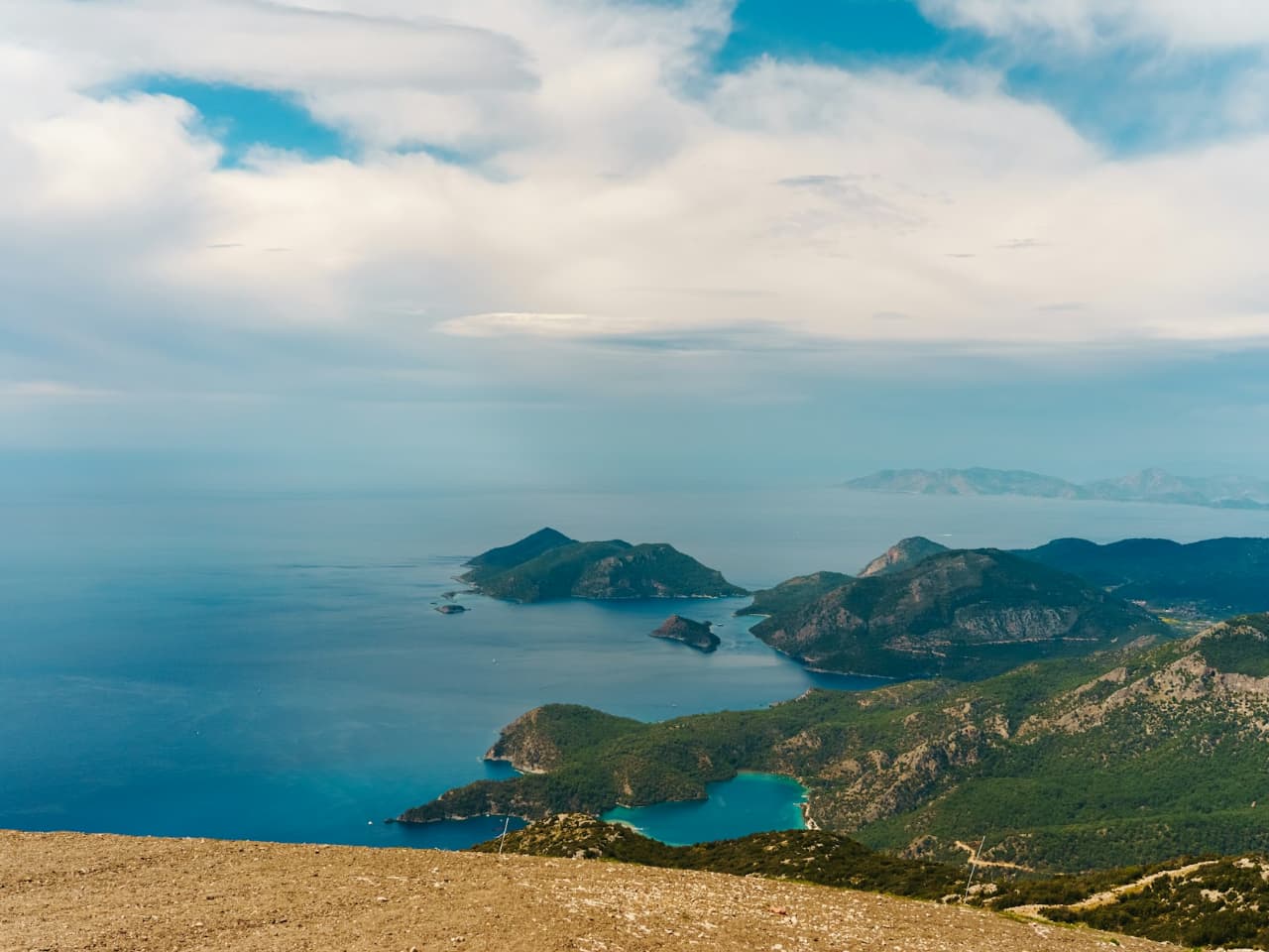Aegean landscape with clear blue waters, gentle mountain curves, and a light blue sky.